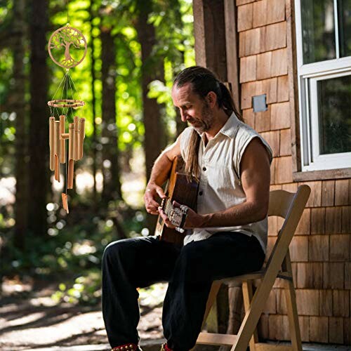 Man playing guitar outdoors near wooden cabin with wind chimes.